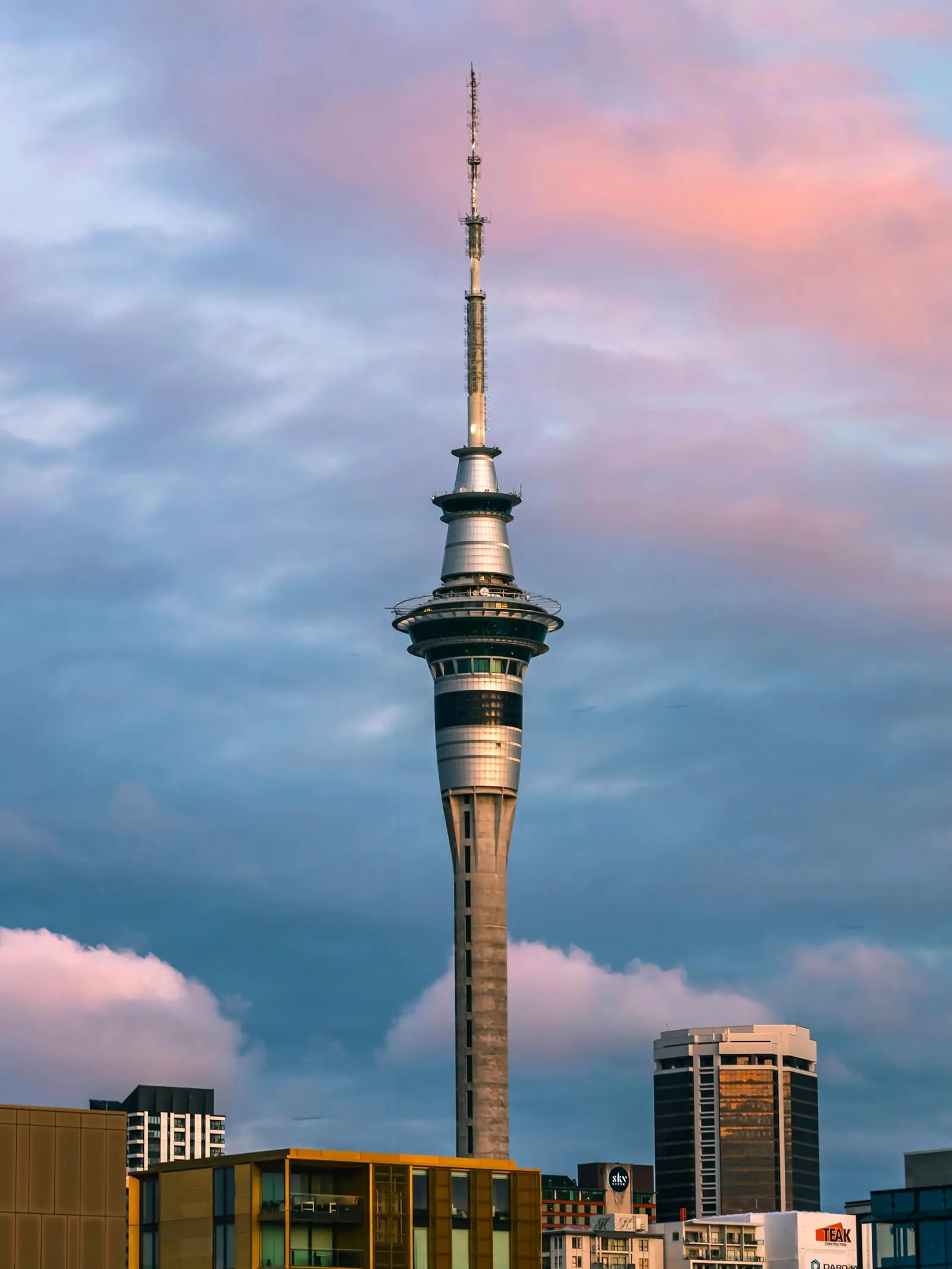 The Sky Tower against pink and blue sunset clouds, rising above Auckland CBD rooftops and a SkyCity sign